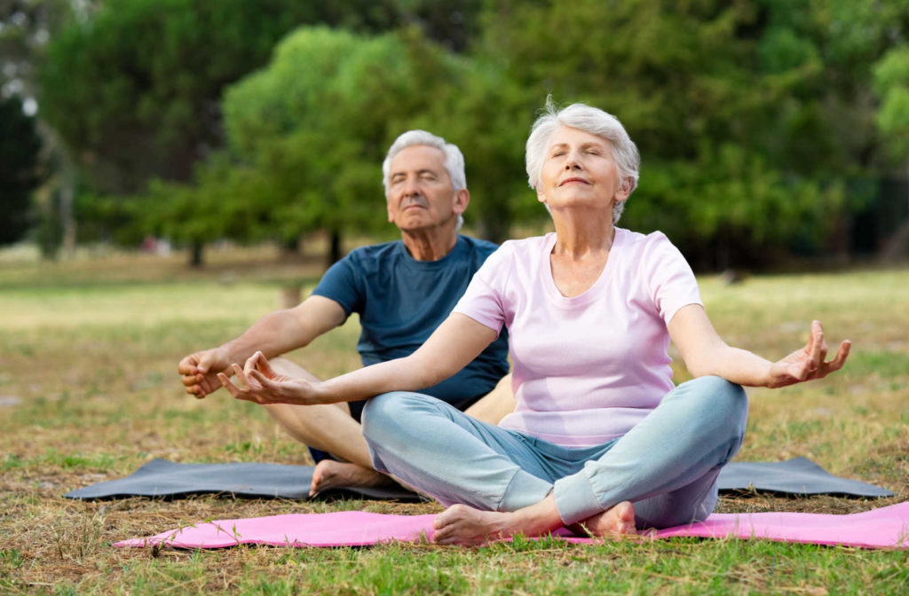 A senior couple meditating on yoga mats outside on the grass.