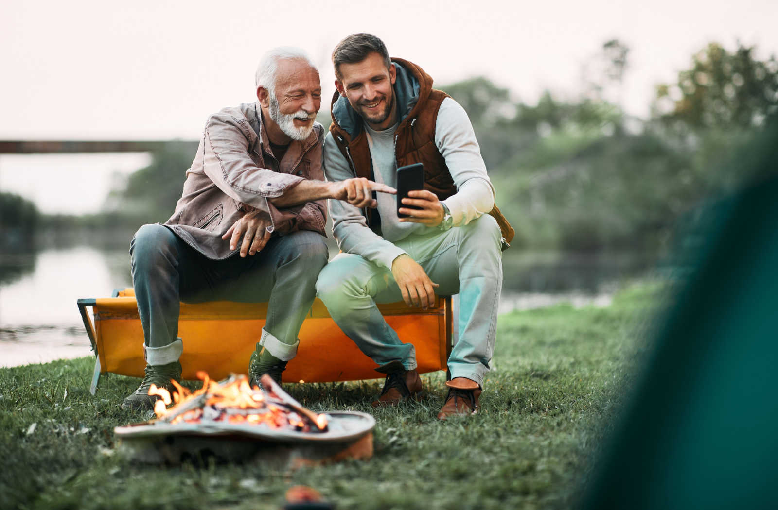 A young man and his elderly father sitting by the campfire at sunset having fun while using a smartphone.