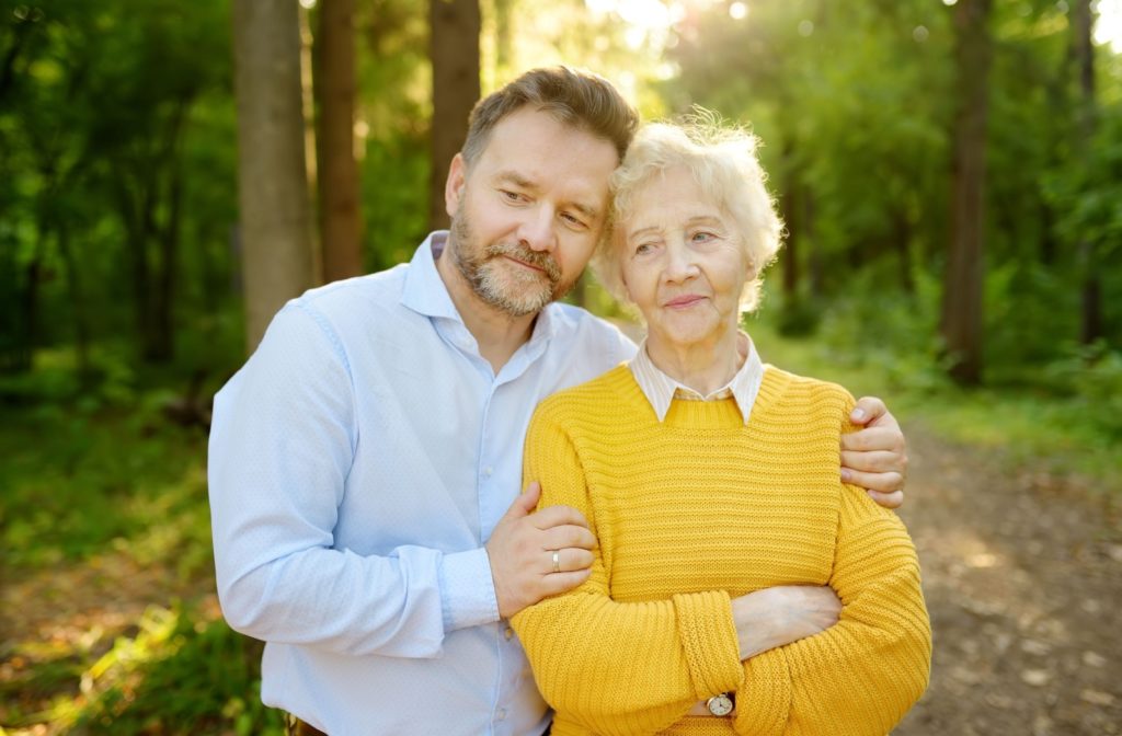 Middle aged man in light blue shirt with arms around and leaning slightly into senior woman in yellow sweater whose arms are crossed.