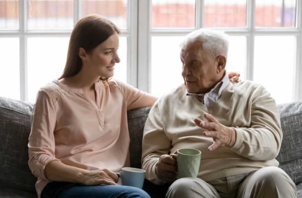 An adult sits on a couch with their older parent, drinking tea while listening to their parent’s concerns about senior living