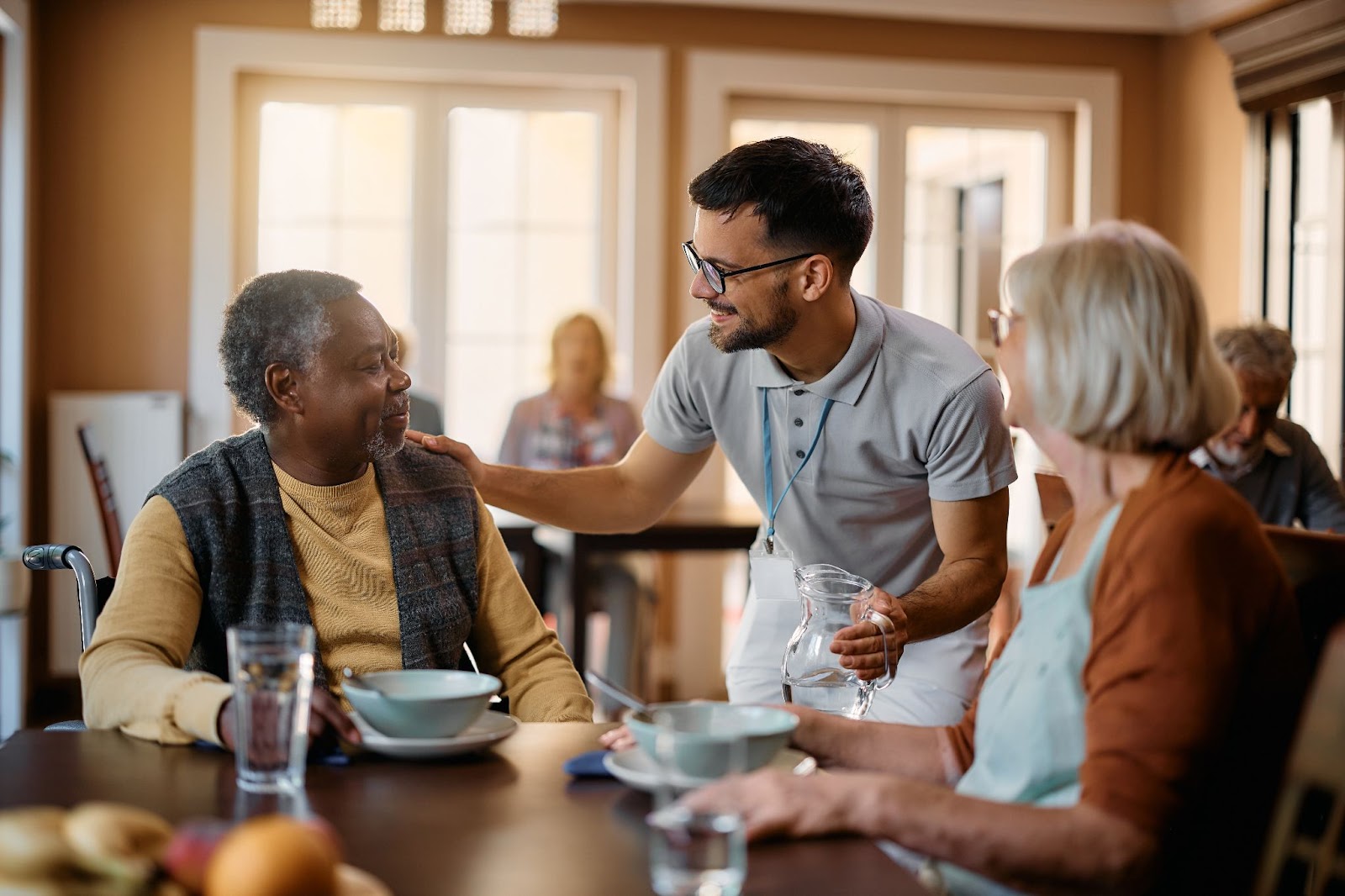 Two seniors sit at a dining room table, having just finished a meal. A server is chatting with them and offering to refill their water glasses. All three have large smiles, clearly enjoying themselves.