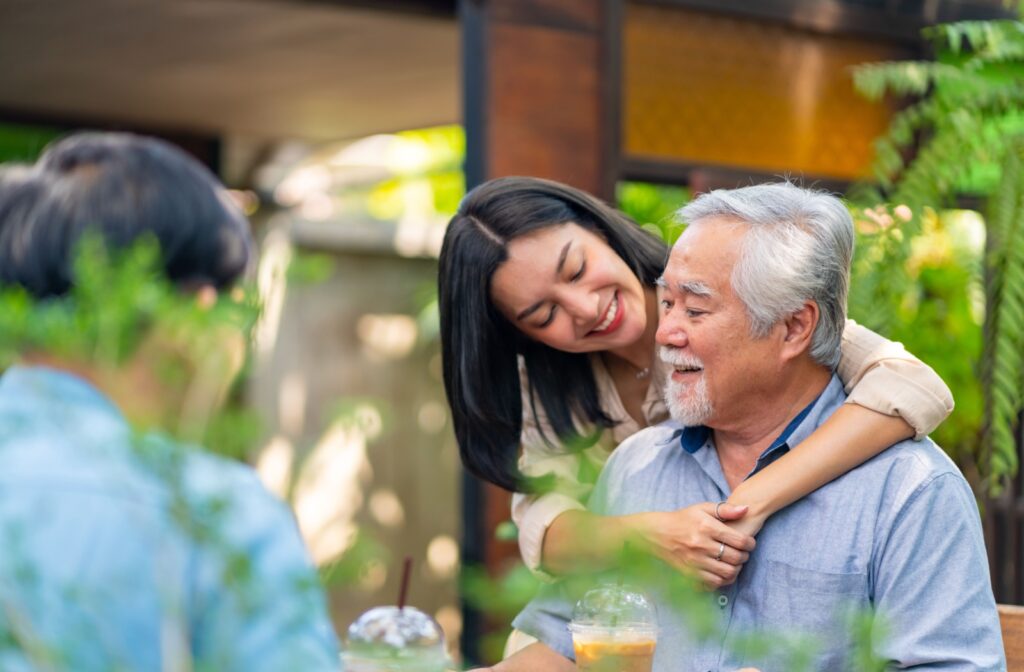 Smiling adult woman with black hair standing behind sitting elderly man with beard with arm around his shoulders.