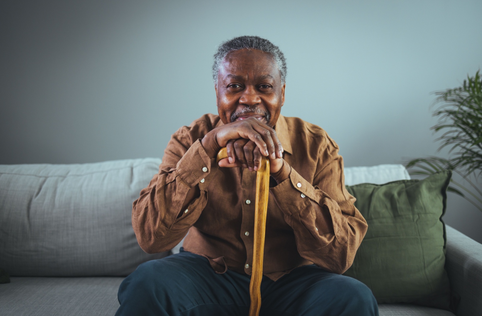 An older adult man sitting on a couch, smiling, and holding a cane.