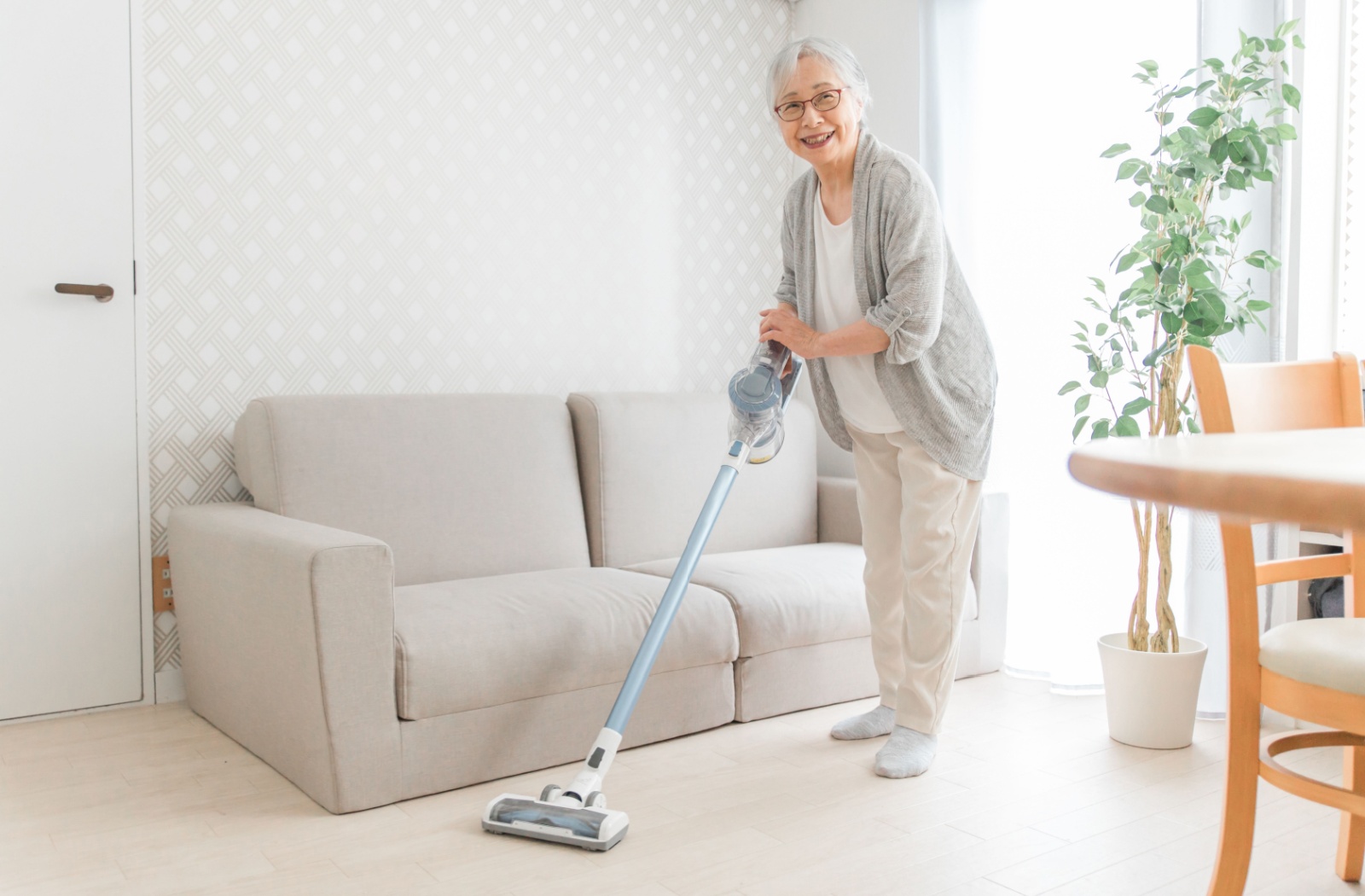 Older adult smiling while vacuuming the living room floor with a lightweight cordless vacuum.