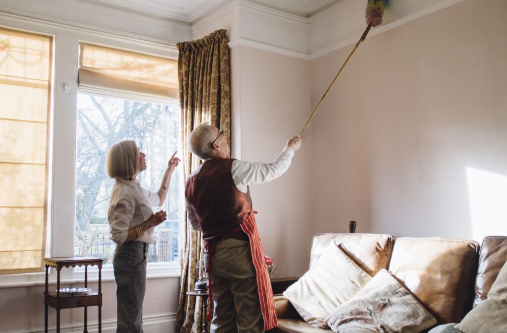 Two people cleaning a living room while one uses an extended duster to reach high corners near the ceiling.