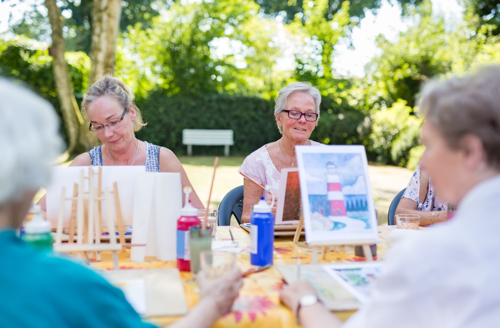 Group of older adults painting at easels outdoors on a sunny day during a creative group activity.