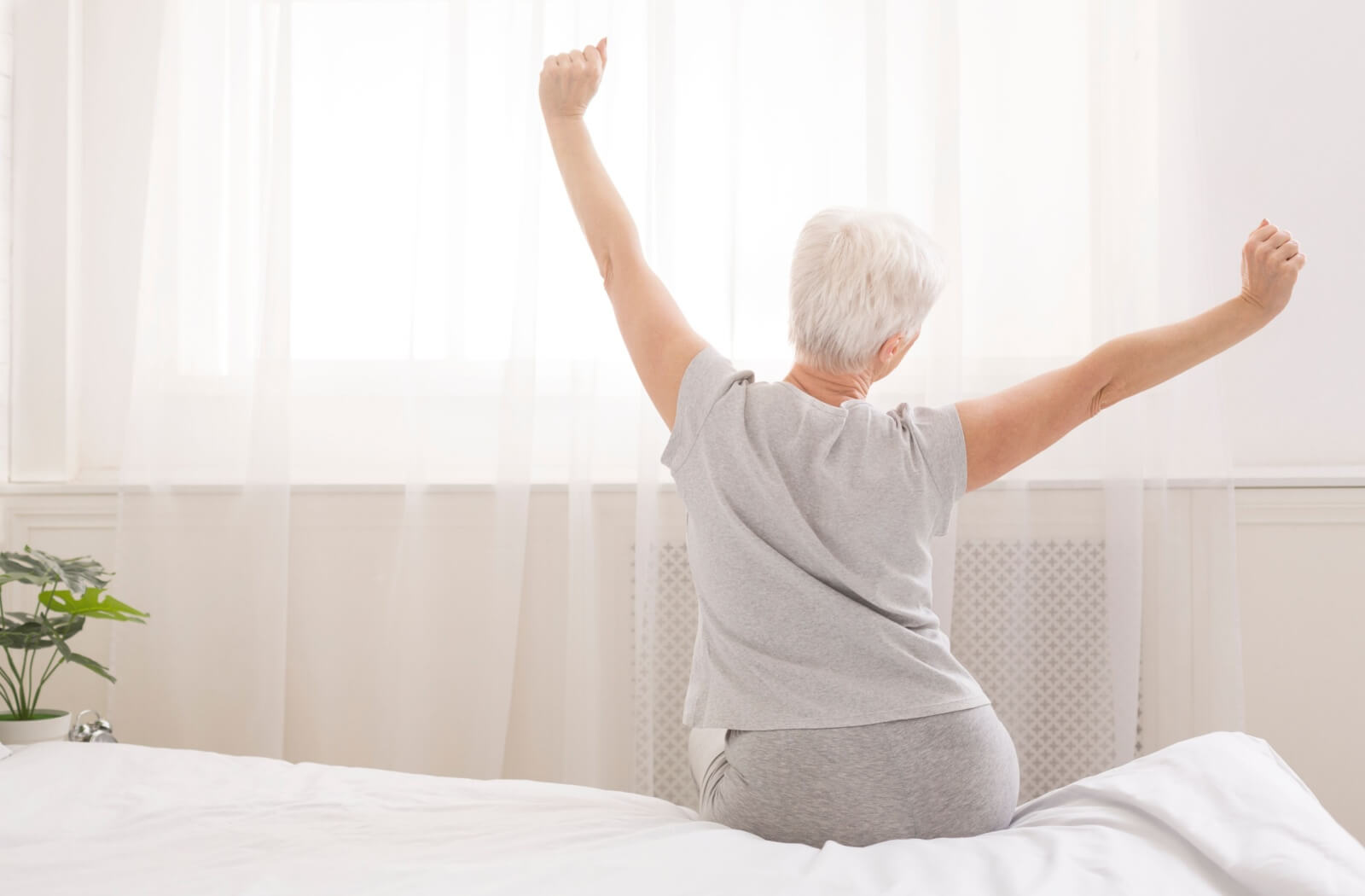 An older adult stretching in the morning in front of the sunlit window of their bedroom.