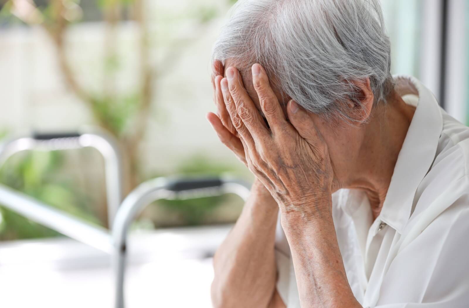 An older adult sits on a bench in a sunlit courtyard and rubs their eyes due to extreme fatigue.