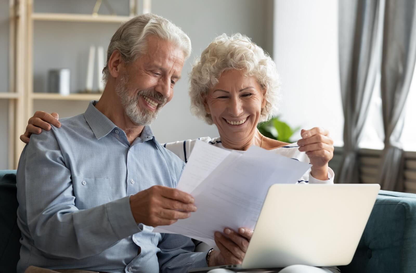 An older couple smiles after printing out their finished application for independent living while sitting arm-in-arm on their couch at home
